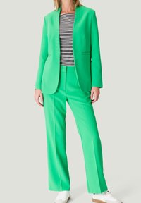 Donna che indossa un completo verde brillante con giacca su misura e pantaloni, abbinato a una maglietta a righe bianche e nere e sneakers bianche.