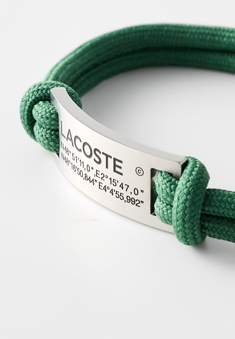Grünes gewobenes Armband mit einem metallischen silbernen Verschluss, der mit "LACOSTE" und geografischen Koordinaten graviert ist. Strukturiertes Gewebe und schlankes Design.