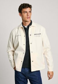 Svetlosivý denimový kabát s cvokmi, s logom a textom "Pepe Jeans". Nosený cez čiernu polo košeľu, v kombinácii s tmavomodrými džínsami.