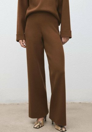 Braune, weit geschnittene Hose kombiniert mit hochhackigen Stiefeletten in Schlangenlederoptik an einer Person, die vor einer schlichten Wand steht.