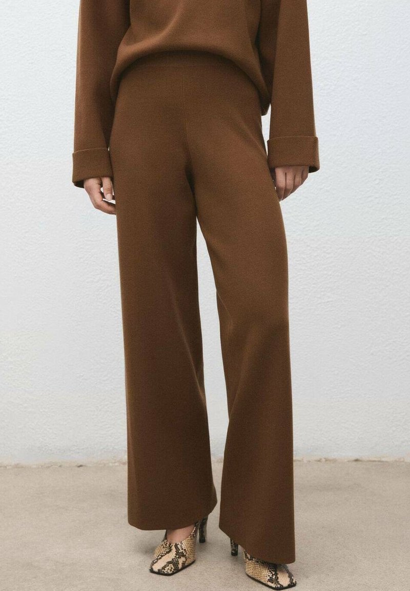 Pantalon marron à jambes larges associé à des bottines à talons hauts avec motif peau de serpent, porté par une personne debout contre un mur uni.