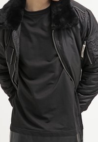 Giacca bomber nera con colletto in pelo sintetico, apertura frontale con zip e tasche laterali. Presenta una finitura texturizzata e un tessuto liscio all'interno.