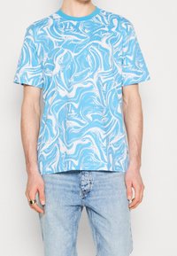 Uomo che indossa una camicia a maniche corte con un motivo swirl blu chiaro e bianco, jeans blu chiaro e un anello d'argento sulla mano destra.