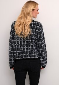 Melnā un baltā kvadrātveida svītra džemperis ar īsu dizainu, adīta tekstūra un ribota apakšmala. Valkāts kopā ar melniem cieši pieguļošiem džinsiem.