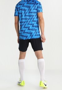 Camiseta deportiva azul y negra con un patrón en zigzag, combinada con pantalones cortos negros, medias blancas hasta la rodilla y zapatillas de fútbol en neón amarillo.