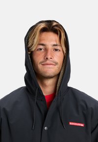 Černá bunda s kapucí, s fleecovou podšívkou a zapínáním na knoflíky. Na hrudi je červené logo Quiksilver. Hladký povrch a ležérní design.