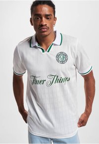 Mann trägt weißes Poloshirt mit grünem Besatz, "Finer Things" Schriftzug auf der Vorderseite und rundem Logo auf der Brust, steht vor weißem Hintergrund.