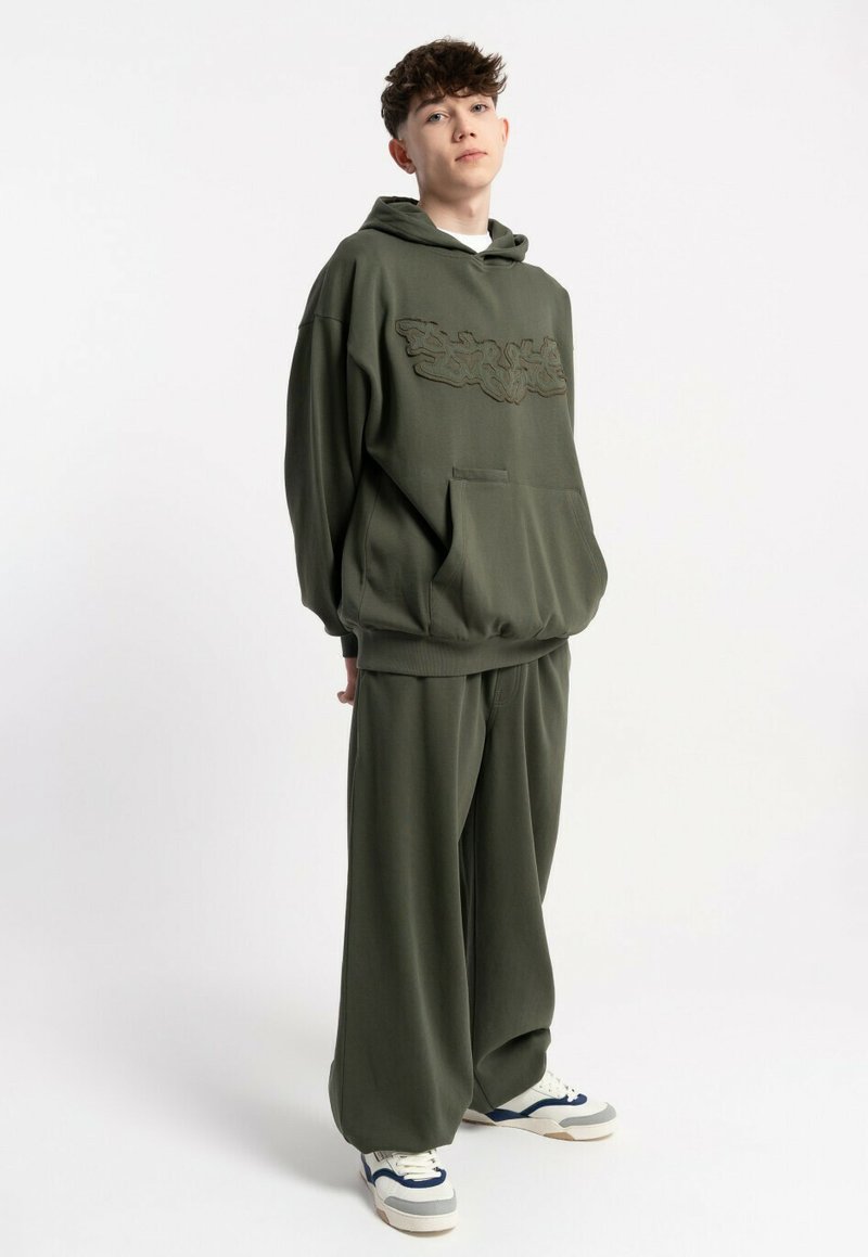 Młoda osoba z krótkimi, kręconymi włosami, nosząca za dużą oliwkowo-zieloną bluzę i dopasowane szerokie spodnie, stojąca z rękami złożonymi za plecami.