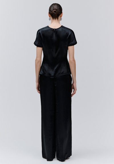 Femme aux cheveux relevés en chignon, vêtue d'un chemisier noir à manches courtes et d'un pantalon à jambes larges, debout de dos sur un fond uni.