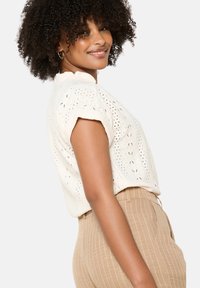 Kreminės spalvos trumpomis rankovėmis bluzonai su aklėmis detalėmis ir banguotu apykaklės kraštu, derinama su šviesiai rudos spalvos dryžuotomis aukštai iki liemens kelnėmis.