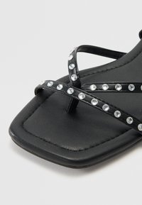Sandale plate noire avec de fines lanières croisées ornées de strass transparents. Bout carré, semelle texturée et matériau lisse ressemblant à du cuir.