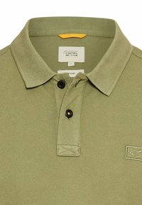 Olivengrøn polo skjorte lavet af økologisk bomuld. Den har en klassisk krave, tre sorte knapper og et lille logo på venstre side.