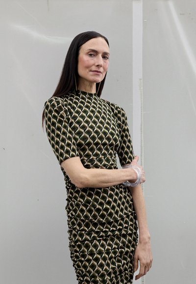 Dopasowana sukienka z geometrycznym wzorem w kolorach zielonym, czarnym i beżowym. Tkanina o fakturze z marszczeniami i krótkimi rękawami. Przezroczysta bransoletka na nadgarstku.