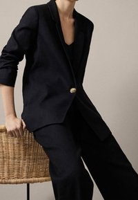 Marineblazer mit tailliertem Schnitt, Ein-Knopf-Verschluss und Revers mit Kerbe; kombiniert mit lockeren, dunklen Hosen; lässiger gewebter Stuhl.
