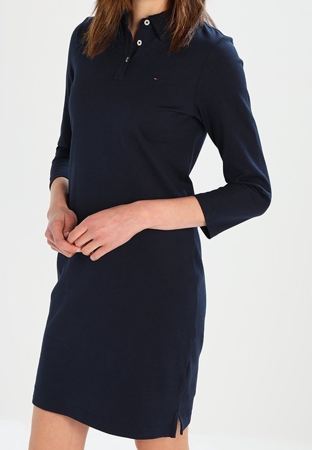 Femme portant une robe polo bleu marine à manches longues avec une petite fente latérale, debout, les mains jointes devant elle.