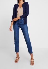 Marinblå cardigan över en beige topp, matchad med högmidjade, raka jeans. Modellen bär lätta, strapiga klackar.