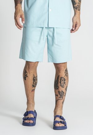 Uomo con pantaloncini azzurri chiari e camicia abbinata, con tatuaggi sulle gambe, che indossa sandali blu con fibbia, in piedi su sfondo semplice.