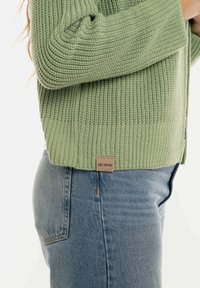 Lichtgroene gebreide cardigan met ribbeltextuur; voorzien van een geweven label aan de zoom. Gecombineerd met hoge blauwe jeans.