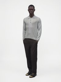 Homme portant un polo gris clair à manches longues, un pantalon noir et des chaussures noires avec embouts dorés, debout les mains dans les poches.