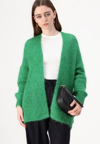 Grønn, fluffy cardigan med åpen front og lange ermer, kombinert med en hvit topp og svart veske. Strikket stoff med tekstur, avslappet passform.