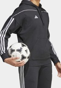 Sudadera negra con acentos blancos, que presenta un bolsillo frontal y puños acanalados. La persona sostiene un balón de fútbol en blanco y negro con una marca.