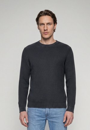 Jeune homme aux cheveux châtain clair, portant un pull côtelé gris foncé et un jean bleu clair, debout devant un fond clair uni.