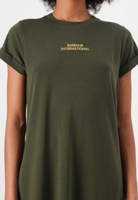 Oliwkowa koszulka z miękkiej bawełny, z podwiniętymi rękawami i haftowanym logo "BARBOUR INTERNATIONAL" w kolorze złotym. Gładka tekstura.