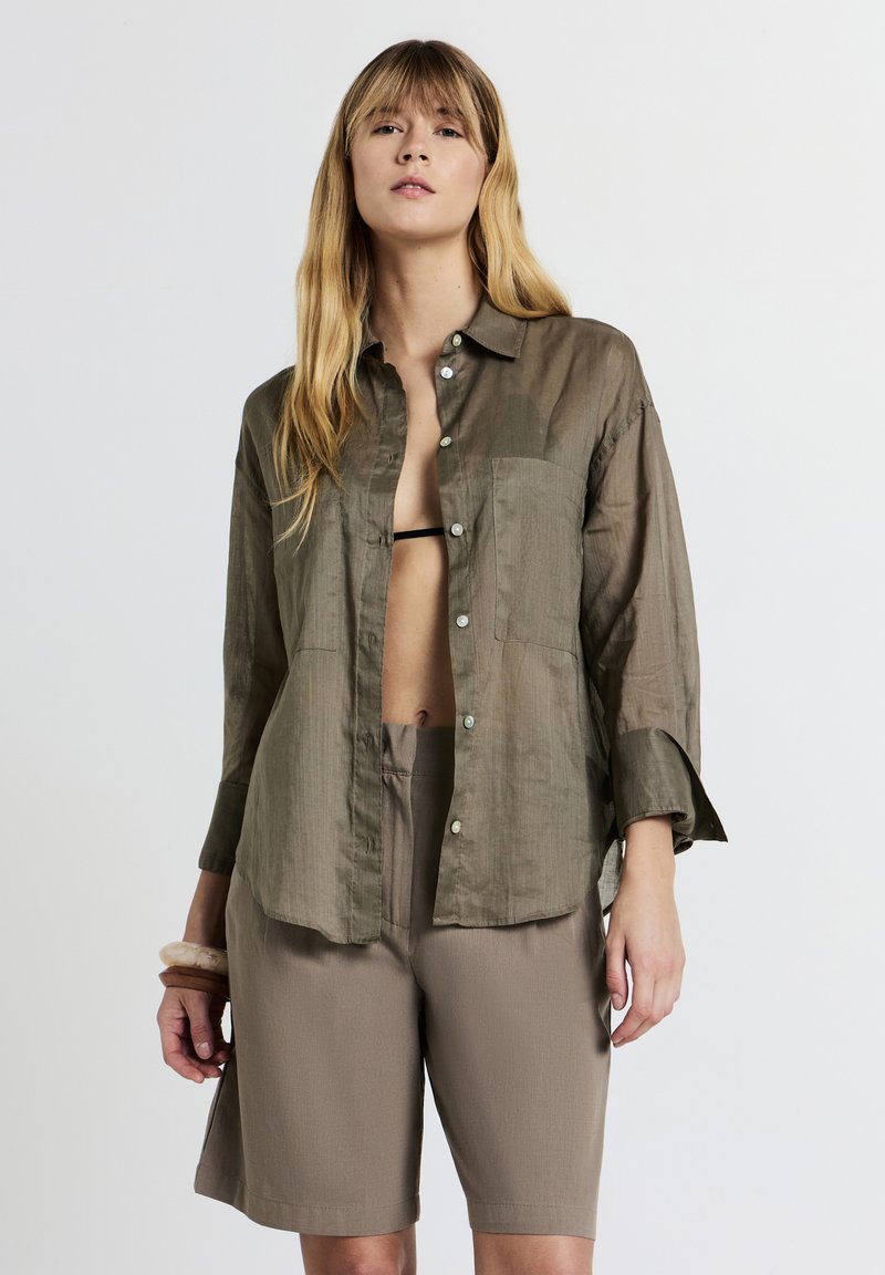 Giovane donna che indossa una camicia trasparente olive green con bottoni sopra un bralette nero e pantaloni corti taupe a vita alta, in piedi su uno sfondo semplice.