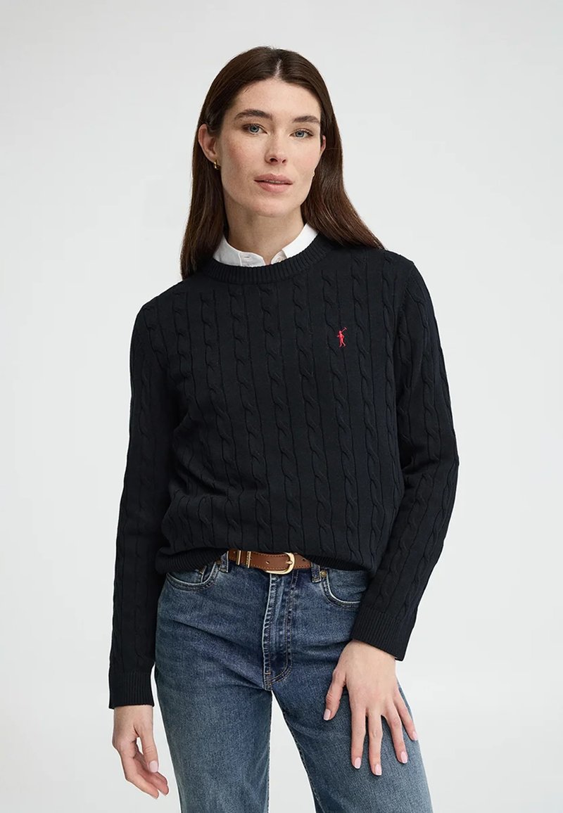 Donna indossa un maglione nero a trecce con logo rosso, camicia bianca con colletto sotto, jeans blu e cintura marrone, in piedi davanti a uno sfondo semplice.