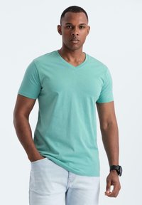 Uomo che indossa una T-shirt verde chiaro a V e jeans azzurri, con una mano in tasca e un orologio nero sull'altro polso.