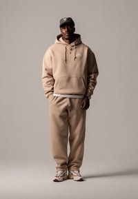 Beige bomull hettegenser med frontlomme og snor, paired med matchende joggebukser. Hvit logodetalj. Røde og hvite sneakers.