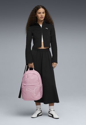 Roze Puma rugzak met een voorvak en logo, gedragen met een zwart cropped jack en een lange rok. Witte sneakers maken de outfit compleet.