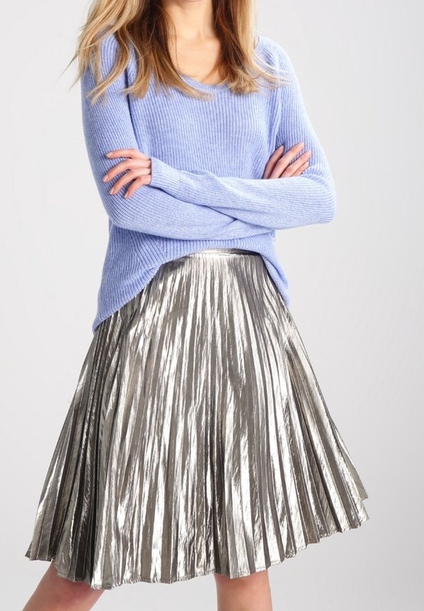 Femme portant un pull en maille violet clair et une jupe midi plissée argentée brillante, les bras croisés.