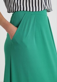 Zelená skládací sukně z tkané látky, se bočními kapsami a hladkou texturou. V kombinaci s černobílým pruhovaným topem.