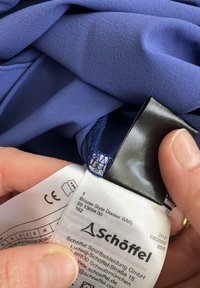 Blue polyester blouse with a smooth texture, featuring a black label with care instructions and a decorative stitching detail on the hem.