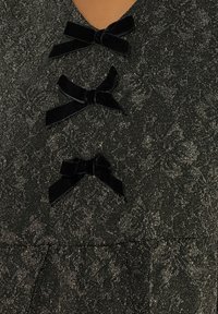 Abito nero con una stoffa texture e luccicante caratterizzata da un motivo floreale. Tre fiocchi in velluto nero adornano la parte anteriore, aggiungendo un tocco di dettaglio.