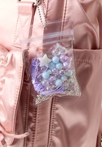 Kirkas muovikassi, jossa on värikkäitä helmiä, naruja ja hopeisia yksityiskohtia, kiinnitetty vaaleanpunaiseen satiinireppuun. Sisältää erilaisia muotoja ja kokoja.