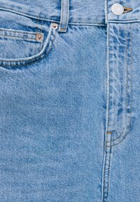 Gros plan sur un jean en denim bleu clair montrant la poche avant, la boucle de ceinture, le bouton et les détails visibles des coutures.