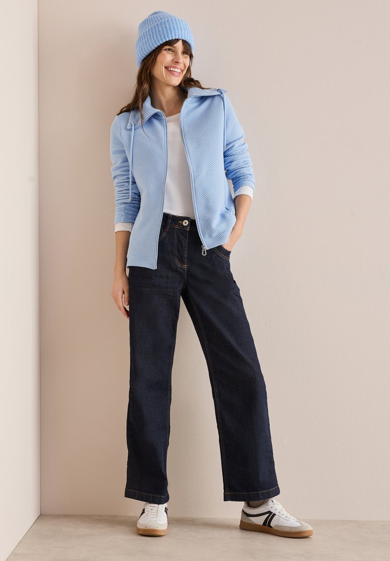 Giacca zip-up azzurra chiara in tessuto texturizzato, t-shirt bianca, jeans larghi scuri e sneakers bianche con dettagli neri, abbinate a un berretto di maglia blu.