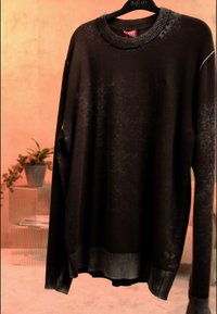 Ciemnobrązowy sweter z długim rękawem, z prążkowanym kołnierzem i mankietami, wiszący na czarnym wieszaku na tle brzoskwiniowej ściany z roślinami w tle.