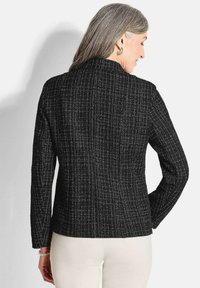 Frau mit grauem Haar, die eine schwarze und weiße strukturierte Jacke mit langen Ärmeln und helle Hosen trägt, von hinten vor einem weißen Hintergrund gezeigt.