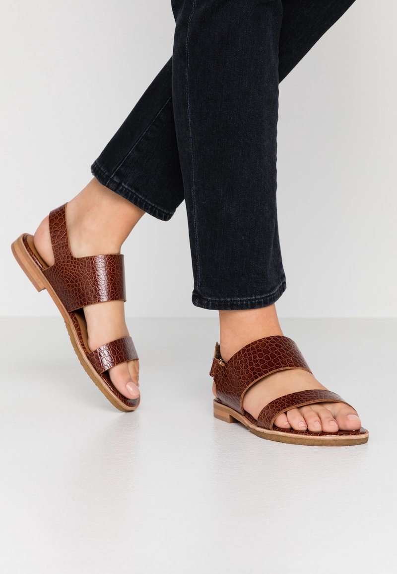 Personne portant un jean foncé et des sandales en cuir marron avec des brides texturées, debout sur un sol clair.