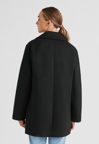 Schwarzer übergroßer Mantel mit Reverskragen, geradem Schnitt und langen Ärmeln. Glatte Textur, minimalistische Details und eine saubere Verarbeitung.