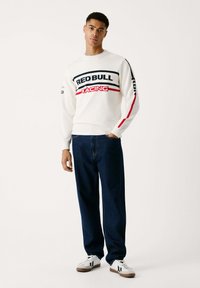 Sudadera blanca con "RED BULL RACING" en negro y rojo, combinada con unos pantalones vaqueros sueltos de color azul oscuro y zapatillas blancas.