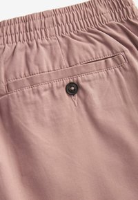 Gros plan sur un pantalon rose à taille élastique avec une poche passepoilée arrière sécurisée par un bouton noir.