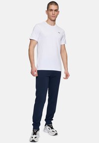 Witte T-shirt met korte mouwen en een klein zwart logo, gecombineerd met marineblauwe joggers en zwart/witte sneakers, ontworpen voor casual gebruik.