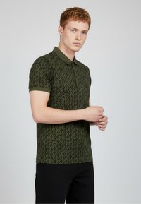 Zelené polo tričko s texturovaným cikcak vzorem, krátkými rukávy a límečkem. Černé detaily na hrudníku. Nošeno s černými kalhotami.