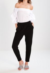 Kvinna i vit avskuren blus med puffärmar, svarta dragsko-byxor och grå slip-on sandaler, stående med händerna i fickorna.