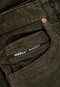 Pantaloni di velluto a coste ampi di colore verde scuro con superficie testurizzata. Presentano un'etichetta nera visibile con la scritta "REELL BAGGY" e un bottone con logo in rame.