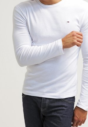 Homme portant une chemise blanche à manches longues avec un petit logo sur la poitrine et un jean foncé, ajustant la manche avec la main droite.